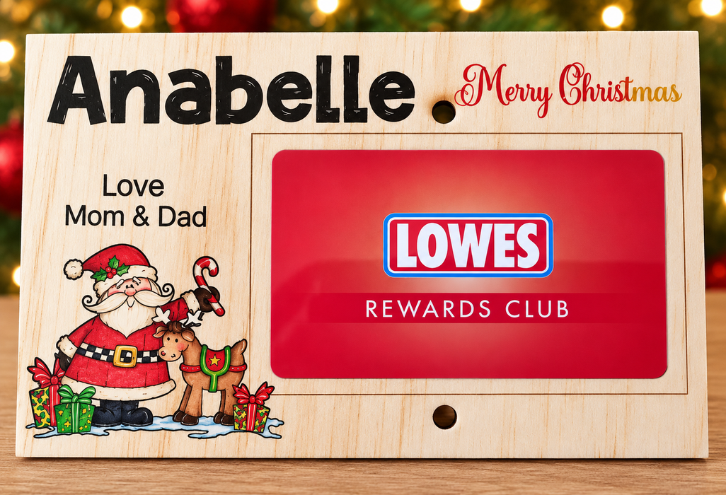 A personalized wooden Christmas photo frame with the names 'Anabelle' and 'Love Mom & Dad', a festive Santa illustration, and a red insert displaying the 'LOWES REWARDS CLUB' logo. The frame is set against a blurred holiday background with warm lights.