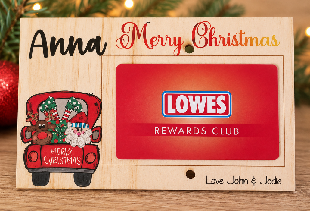 A personalized wooden Christmas card holder plaque engraved with the name 'Anna', 'Merry Christmas', a red truck illustration, and 'Love John & Jodie'. A red Lowes Rewards Club card is inserted into the center slot.