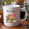 A white ceramic coffee mug with a colorful cartoon graphic and text, displayed on a wooden surface with a blurred background of plants and dried flowers.
