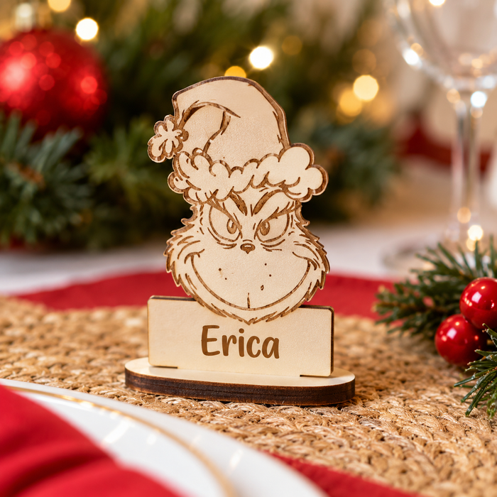 Personalized laser-cut wooden Grinch place card holder with a Santa hat and the name 'Erica' engraved, set on a woven placemat with a blurred Christmas background.