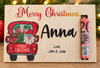 A personalized wooden Christmas plaque on a tabletop with blurred holiday lights in the background. The light natural wood plaque features a red hand-painted truck carrying Christmas gifts and Santa, with the text 'Merry Christmas', the name 'Anna' in large black script, and 'Love John & Jodie' below.