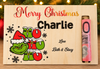 A personalized wooden Christmas gift box engraved with 'Merry Christmas Charlie' and a Grinch-themed 'Ho Ho Ho' design, with a rolled pink twenty-dollar banknote tucked into the side slot. The box is set against a festive background with blurred Christmas lights and greenery.