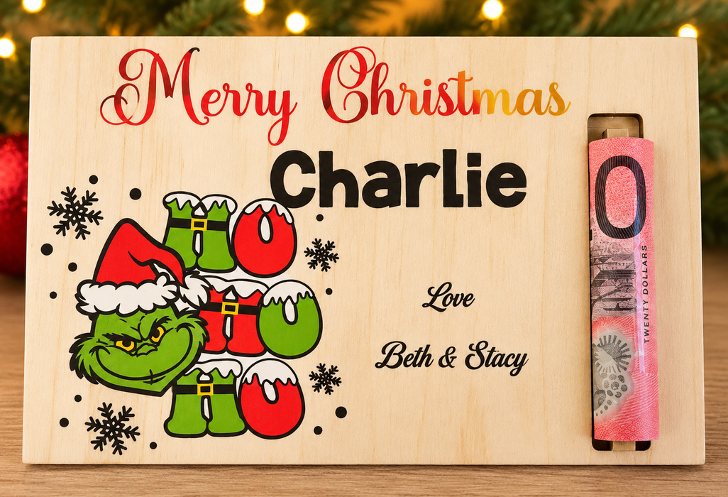 A personalized wooden Christmas gift box engraved with 'Merry Christmas Charlie' and a Grinch-themed 'Ho Ho Ho' design, with a rolled pink twenty-dollar banknote tucked into the side slot. The box is set against a festive background with blurred Christmas lights and greenery.