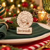 A personalized wooden place card stand engraved with a Christmas tree and the name 'Jeremiah', displayed on a dark green napkin over a white plate in a festive holiday setting with blurred lights and ornaments.