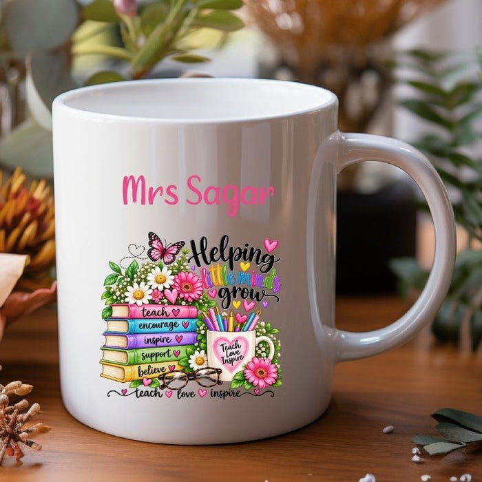 A white ceramic mug with a colorful teacher-themed graphic and the text 'Mrs Sagar' and 'Helping students grow,' shown on a wooden surface with blurred plants in the background.