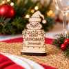 A personalized wooden Grinch sign with engraved text reading 'MERRY GRINCHMAS Jeremiah' stands on a woven placemat in a festive holiday setting with blurred Christmas lights, greenery, and red ornaments in the background.