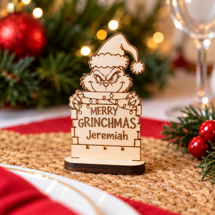A personalized wooden Grinch sign with engraved text reading 'MERRY GRINCHMAS Jeremiah' stands on a woven placemat in a festive holiday setting with blurred Christmas lights, greenery, and red ornaments in the background.