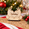 A personalized wooden bunny place card with a heart-shaped center and the name 'Nicholas' engraved, set on a woven placemat with a blurred Christmas background.