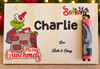 Personalized Christmas-themed wooden plaque featuring a Grinch illustration, the name Charlie, and a small vertical slot holding a pink-and-gray decorative rod. The plaque also reads Merry Grinchmas and Love Beth & Stacy, with blurred holiday lights in the background.