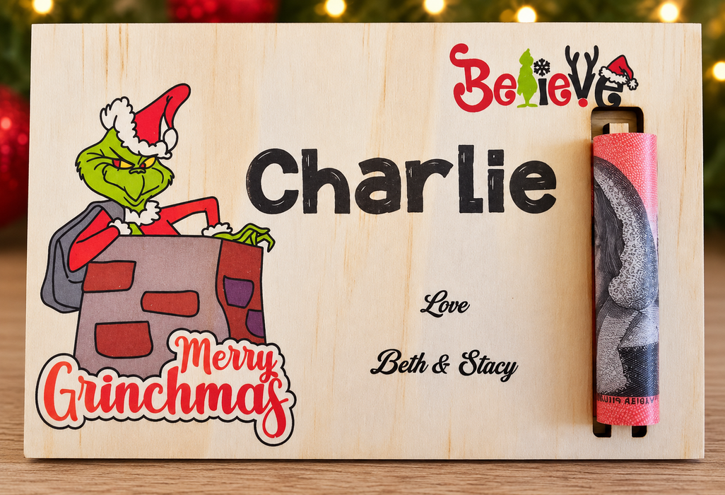 Personalized Christmas-themed wooden plaque featuring a Grinch illustration, the name Charlie, and a small vertical slot holding a pink-and-gray decorative rod. The plaque also reads Merry Grinchmas and Love Beth & Stacy, with blurred holiday lights in the background.