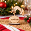 A personalized wooden Christmas ornament place card stand featuring a laser-cut hand reaching down to a nameplate engraved with 'Lewinton'. The stand is displayed on a woven placemat with a red tablecloth, pine branches, and festive lights in the background.