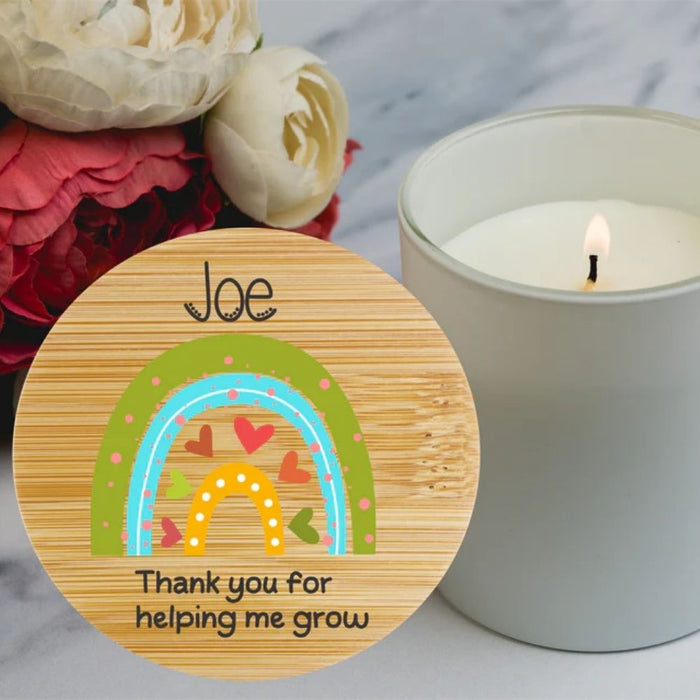 A round personalized bamboo candle sticker with a colourful rainbow design and personalised is shown next to a lit white candle on a marble surface with flowers in the background.