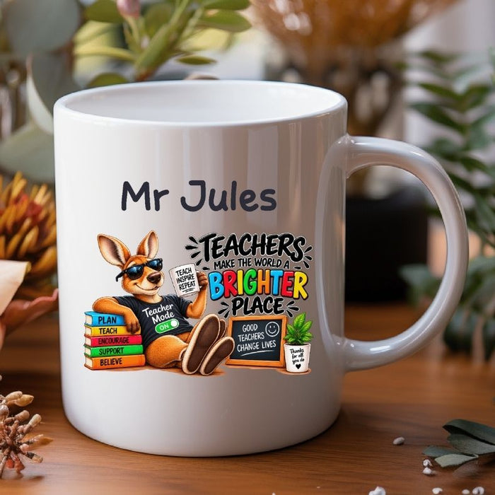 A white ceramic mug with a custom printed design featuring a cartoon kangaroo and the text 'Mr Jules TEACHERS MAKE THE WORLD A BRIGHTER PLACE', set on a wooden table with a blurred background of plants and decor.