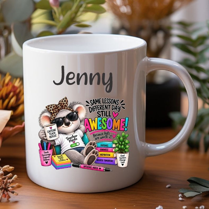 A white ceramic mug with a colorful cartoon illustration of a koala wearing sunglasses and a leopard-print hat, next to the name 'Jenny' in black script. The mug is placed on a wooden surface with blurred plants in the background.