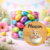 A clear glass jar filled with colorful foil Easter eggs, set on a pastel surface with flowers and bunting in the background. A round wooden-style label overlay in front shows the name Mason and a bunny in a floral egg.