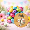 A clear glass jar filled with colorful foil Easter eggs, set on a pastel pink surface with flowers and bunting in the background. A round wooden-style overlay in front shows the name 'Judy' above a cartoon bunny holding a floral egg.