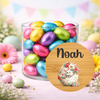 A clear glass jar filled with colorful foil-wrapped chocolate eggs on a pastel floral surface, with a round wooden-style label in front showing the name Noah and a floral bunny illustration.