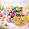 A clear glass jar filled with colorful foil-wrapped chocolate eggs, styled on a pastel surface with flowers and Easter-themed decorations. A round wooden label in front shows the name Olivia and a cartoon bunny.