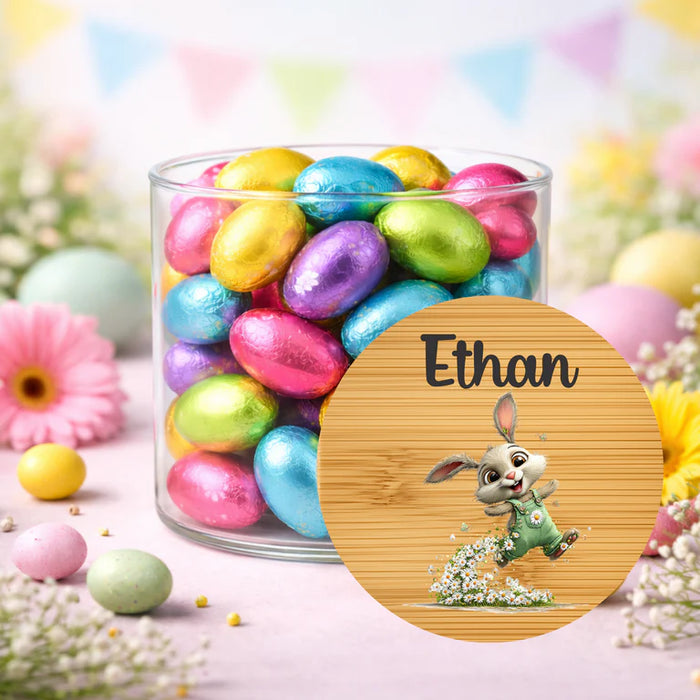 A clear glass jar filled with colorful foil Easter eggs, set on a pastel surface with flowers and bunting in the background. A round wooden-style label in front shows the name Ethan and a cartoon bunny jumping in flowers.