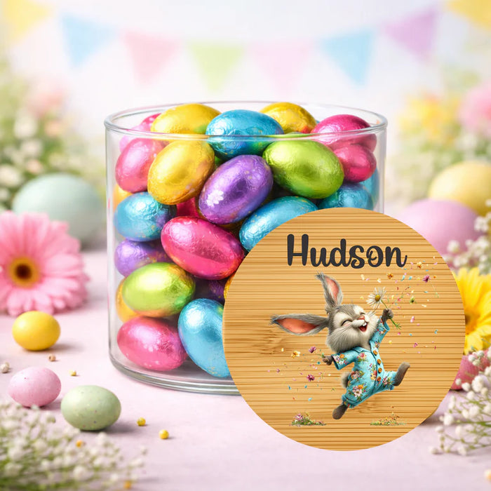 A clear cylindrical glass jar filled with colorful foil Easter eggs, placed on a pastel surface with flowers and bunting in the background. In front of the jar is a round wooden-style label or coaster featuring a cartoon bunny in blue clothing and the name 'Hudson' in black script.