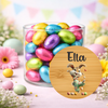 A clear glass jar filled with colorful foil-wrapped Easter eggs, styled on a pastel surface with decorative flowers, eggs, and a round wooden label in front. The label shows the name 'Ella' and a cartoon bunny holding an egg.