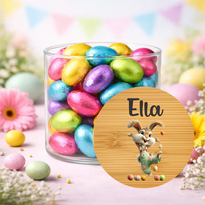 A clear glass jar filled with colorful foil-wrapped Easter eggs, styled on a pastel surface with decorative flowers, eggs, and a round wooden label in front. The label shows the name 'Ella' and a cartoon bunny holding an egg.