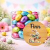 A clear glass jar filled with colorful foil-wrapped chocolate eggs on a pastel Easter-themed background, with a round wooden label in front showing the name Finn and a running bunny illustration.
