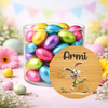 A clear glass jar filled with colorful foil-wrapped chocolate eggs on a pastel Easter-themed background, with a round custom label in front showing the name 'Armi' and a cartoon bunny illustration.