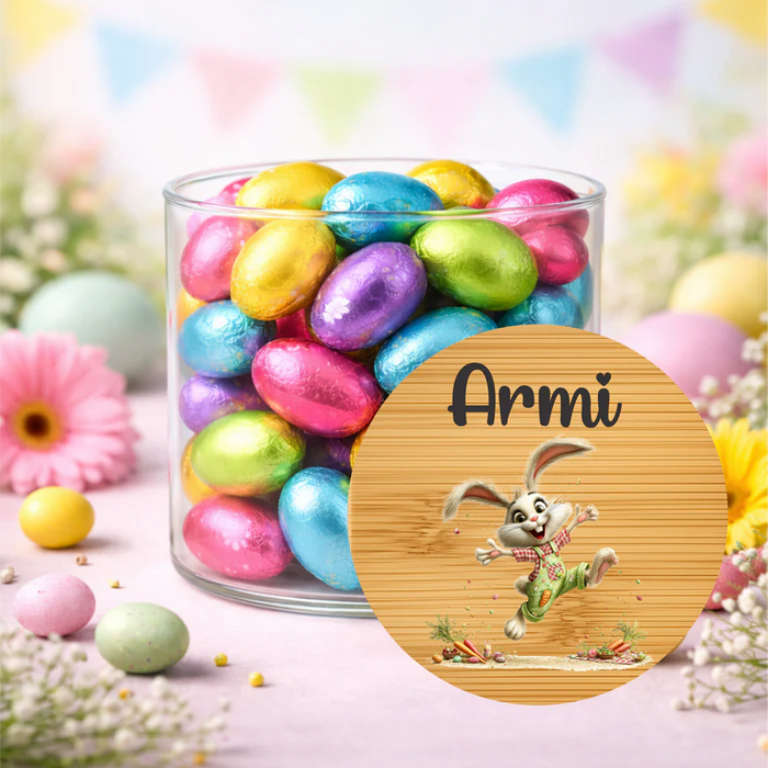 A clear glass jar filled with colorful foil-wrapped chocolate eggs on a pastel Easter-themed background, with a round custom label in front showing the name 'Armi' and a cartoon bunny illustration.