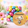 A clear cylindrical glass jar filled with colorful foil-wrapped chocolate eggs, set on a pastel pink surface with decorative flowers and bunting in the background. A round wooden-style label overlay in front shows a cartoon rabbit holding Easter eggs and the name 'Alexander' in black script.