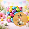 A clear glass jar filled with colorful foil Easter eggs, styled with pastel flowers and eggs on a pink surface. A round wooden-style label in front reads 'Evie' and shows a bunny holding a decorated egg with tulips.