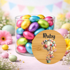 A clear glass jar filled with colorful foil-wrapped chocolate eggs, styled on a pastel surface with flowers and bunting in the background. A round wooden name plaque featuring the name 'Ruby' and a bunny hot air balloon illustration is shown in front.