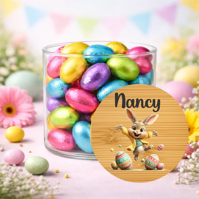 A clear glass jar filled with colorful foil Easter eggs, shown on a pastel floral surface with decorative eggs and flowers around it. A round custom label in front shows the name Nancy above a cartoon bunny with Easter eggs.