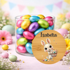 A clear glass jar filled with colorful foil Easter eggs, set on a pastel pink surface with decorative flowers and eggs around it. A round wooden-style label overlay shows the name 'Isabella' above a cartoon bunny holding eggs.