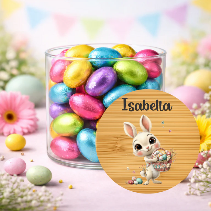 A clear glass jar filled with colorful foil Easter eggs, set on a pastel pink surface with decorative flowers and eggs around it. A round wooden-style label overlay shows the name 'Isabella' above a cartoon bunny holding eggs.