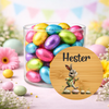 A clear glass jar filled with colorful foil-wrapped Easter eggs, styled on a pastel pink surface with flowers and decorative bunting in the background. A round wooden-style label in front reads 'Hester' and shows a bunny illustration.