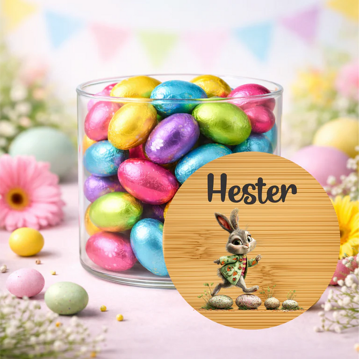 A clear glass jar filled with colorful foil-wrapped Easter eggs, styled on a pastel pink surface with flowers and decorative bunting in the background. A round wooden-style label in front reads 'Hester' and shows a bunny illustration.