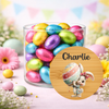 A clear glass jar filled with colorful foil Easter eggs, styled with pastel eggs, flowers, and bunting in a soft spring setting. A round wooden-style label in front shows the name 'Charlie' above a cartoon bunny holding a decorated egg.