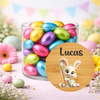 A clear glass jar filled with colorful foil-wrapped Easter eggs, set on a pastel surface with flowers and bunting in the background. A round wooden-style label in front shows a white bunny holding a paintbrush and the name Lucas.