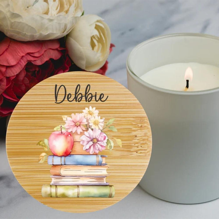 A round wooden coaster or placemat with a printed illustration of stacked books, a red apple, and pink and white flowers, personalised in script at the top. It is styled on a marble surface next to a lit white candle and large red and white flowers.