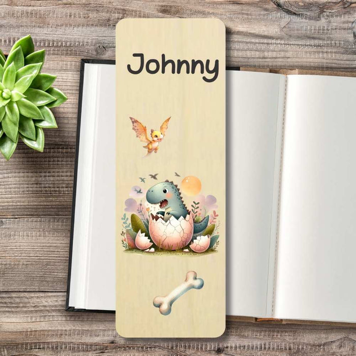A personalized wooden bookmark featuring a cartoon dinosaur hatching from an egg, personalised at the top, and a bone illustration, displayed on a rustic wooden table next to an open notebook and a green succulent.