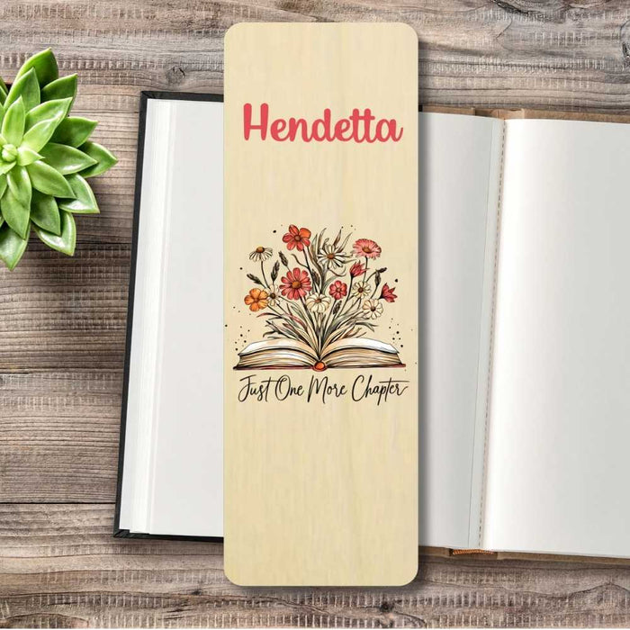 A personalized wooden bookmark personalised in red and an illustration of an open book with flowers, placed on top of an open book on a rustic wood surface with a small succulent.