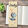 A personalized wooden bookmark featuring a cute cartoon blue dinosaur, a small bird, and personalised at the top, displayed on a rustic wooden tabletop next to an open book and a green succulent.