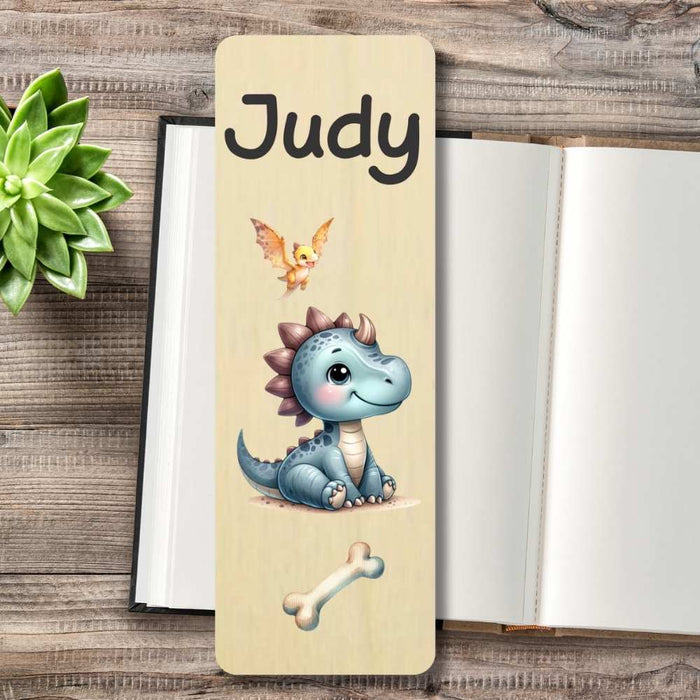 A personalized wooden bookmark featuring a cute cartoon blue dinosaur, a small bird, and personalised at the top, displayed on a rustic wooden tabletop next to an open book and a green succulent.