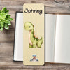 A personalized wooden bookmark featuring a cute green dinosaur illustration printed, styled on an open notebook with a small succulent plant in the corner.