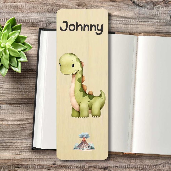 A personalized wooden bookmark featuring a cute green dinosaur illustration printed, styled on an open notebook with a small succulent plant in the corner.