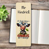 A personalized wooden bookmark styled on an open book with a small green succulent. The bookmark features a cartoon cow wearing glasses and the text 'it's tough being a Genius but I'm married'.