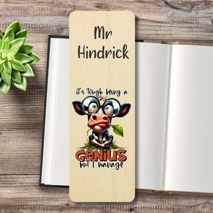 A personalized wooden bookmark styled on an open book with a small green succulent. The bookmark features a cartoon cow wearing glasses and the text 'it's tough being a Genius but I'm married'.