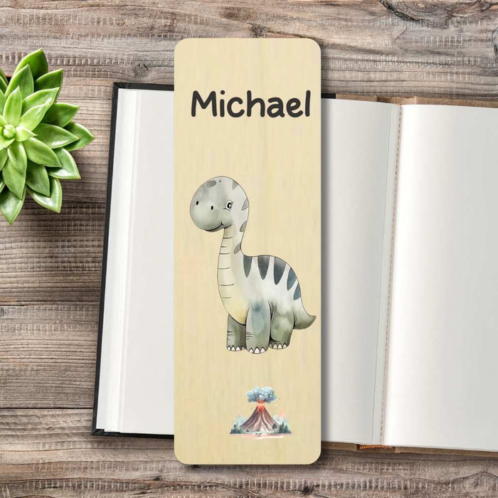 A personalized wooden bookmark featuring a cartoon long-neck dinosaur and the name printed on it, styled on top of an open notebook with a small succulent in the corner.