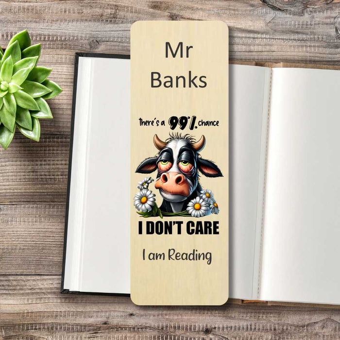 A personalized wooden book bookmark with a cartoon cow and the text 'here's a 99% chance I DON'T CARE I am Reading' is displayed on an open book on a wooden table next to a green succulent.